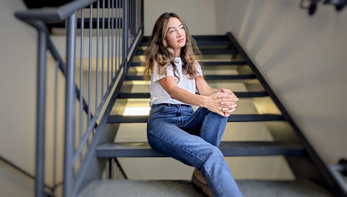 A woman in casual blue jeans and a white t-shirt sits thoughtfully on indoor stairs, pausing with a gentle expression.