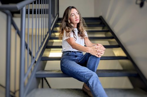A woman in casual blue jeans and a white t-shirt sits thoughtfully on indoor stairs, pausing with a gentle expression.