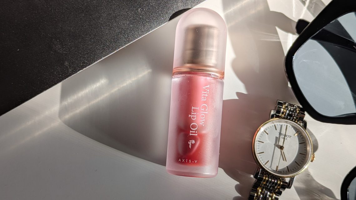 AXIS-Y Vita Glow Lip Oil Chilled Berry in a frosted pink tube placed on a white surface beside a gold Tissot watch and black sunglasses.