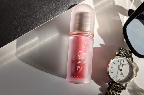 AXIS-Y Vita Glow Lip Oil Chilled Berry in a frosted pink tube placed on a white surface beside a gold Tissot watch and black sunglasses.
