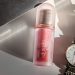 AXIS-Y Vita Glow Lip Oil Chilled Berry in a frosted pink tube placed on a white surface beside a gold Tissot watch and black sunglasses.