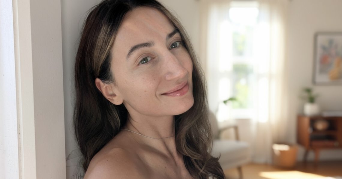 Portrait of a woman with glowing skin without foundation, smiling in soft natural light, with a bright minimal interior background.