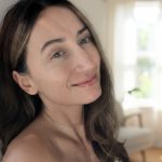 Portrait of a woman with glowing skin without foundation, smiling in soft natural light, with a bright minimal interior background.