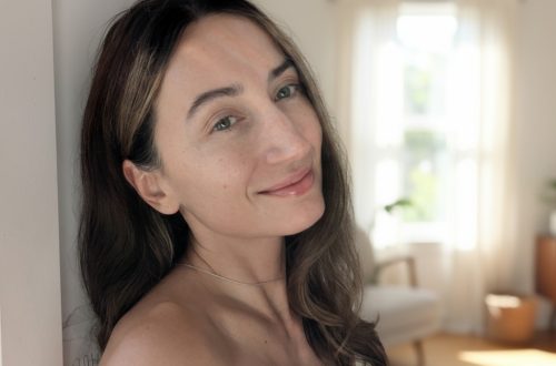 Portrait of a woman with glowing skin without foundation, smiling in soft natural light, with a bright minimal interior background.